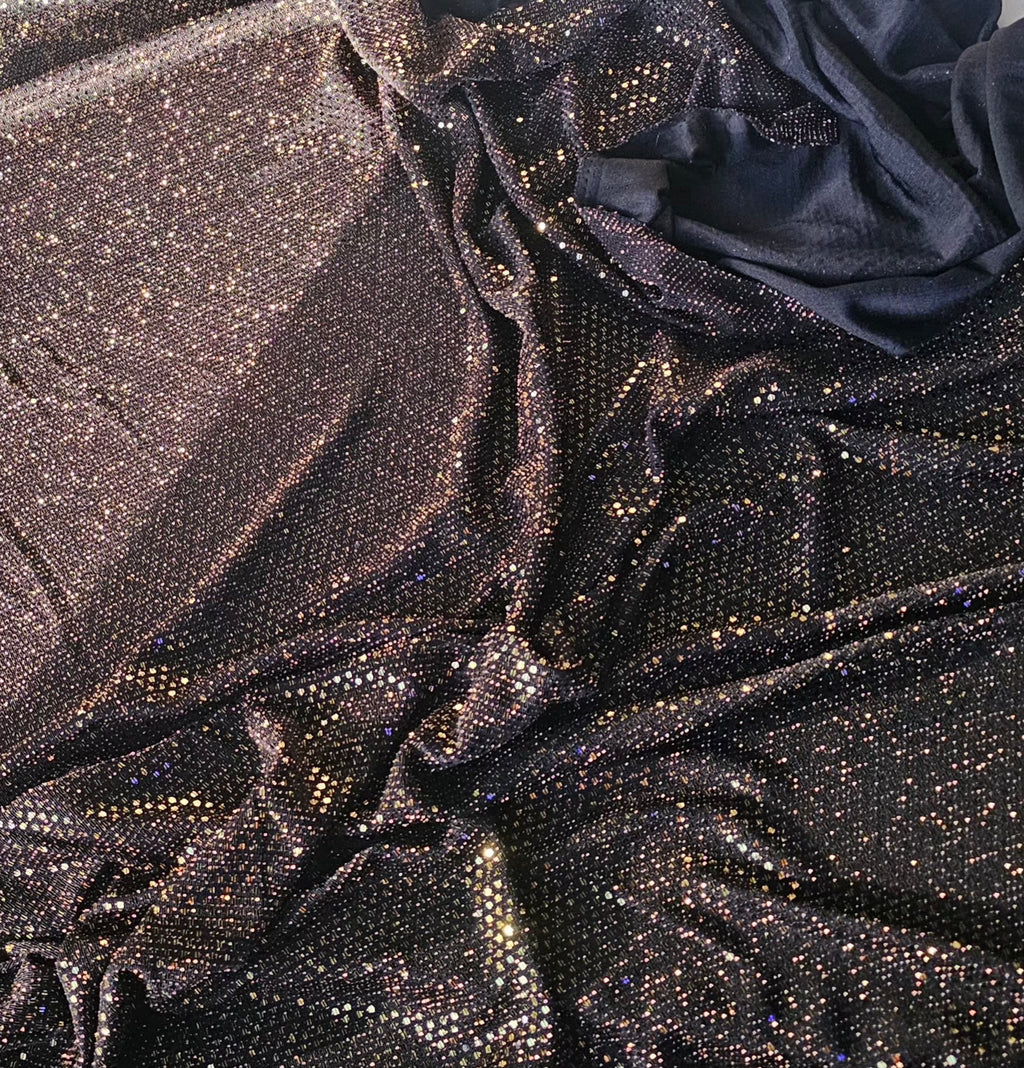 A close-up of draped fabric covered in dense, reflective rhinestones or sequins, showing a dark metallic sheen with scattered warm gold and silver sparkle against a black base.