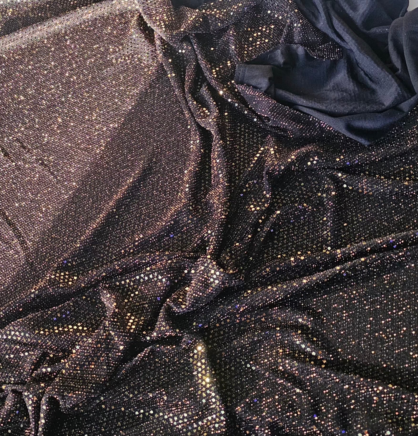 A close-up of draped fabric covered in dense, reflective rhinestones or sequins, showing a dark metallic sheen with scattered warm gold and silver sparkle against a black base.