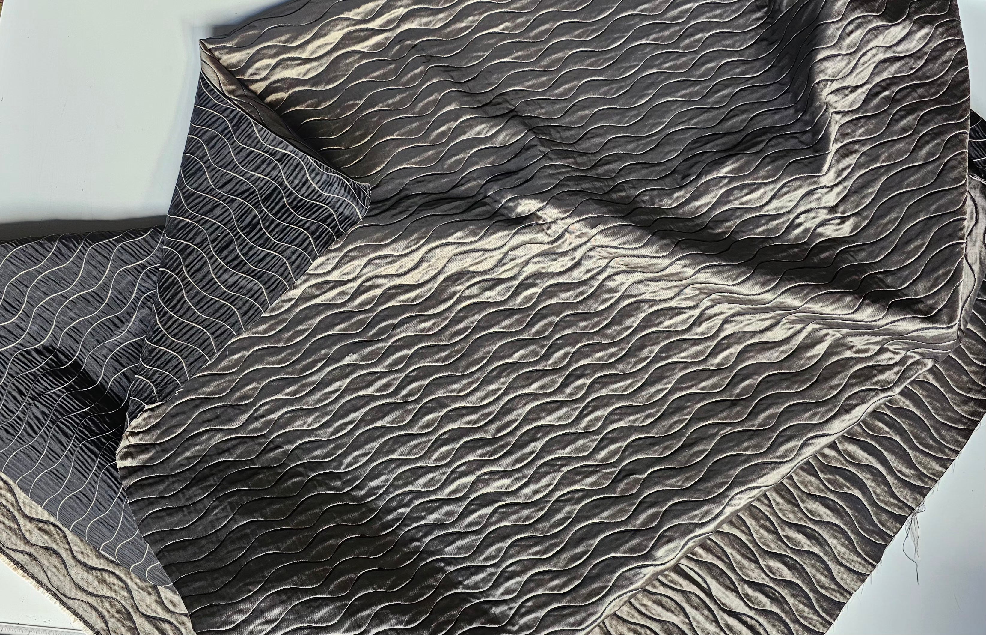 A close-up of a dark gray quilted fabric with a wavy, textured pattern and a matching pillowcase, laid out on a light background.