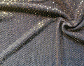 A close-up of shimmering fabric with dense small reflective sequins or rhinestones in a dark metallic gray/silver base, creating bright gold and silver highlights across the surface. The fabric is draped in soft folds.