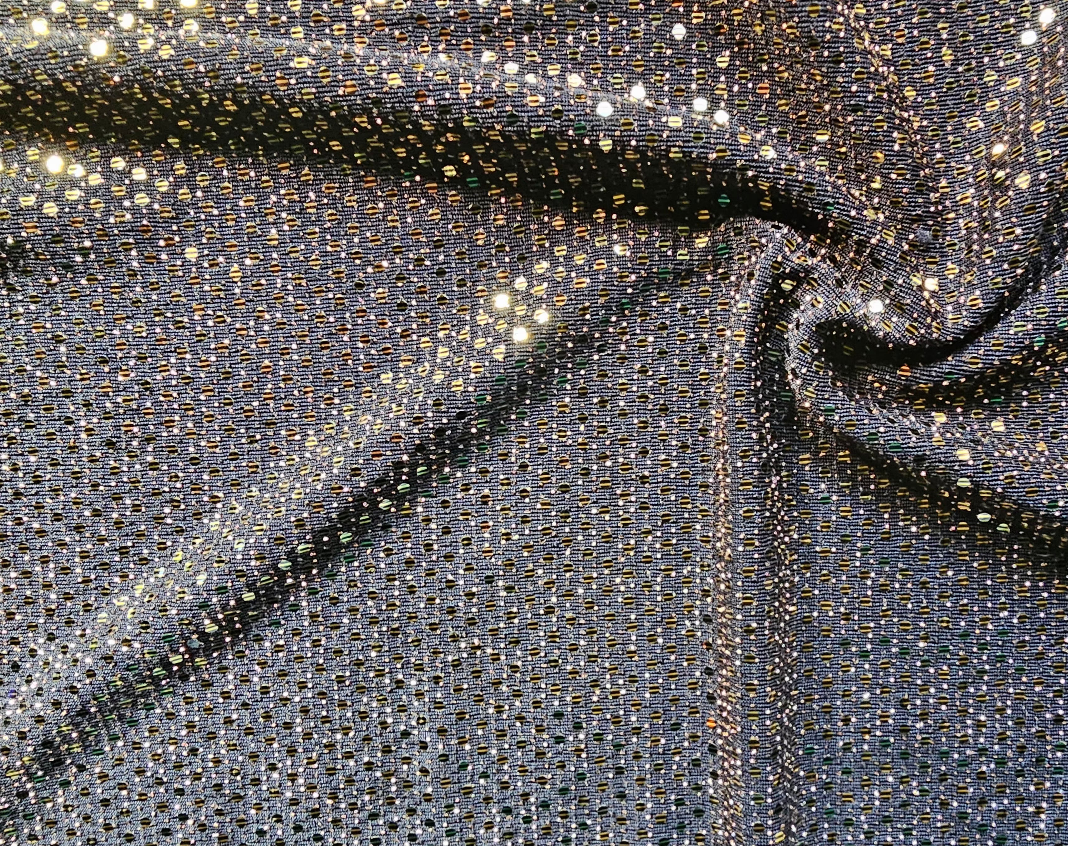 A close-up of shimmering fabric with dense small reflective sequins or rhinestones in a dark metallic gray/silver base, creating bright gold and silver highlights across the surface. The fabric is draped in soft folds.