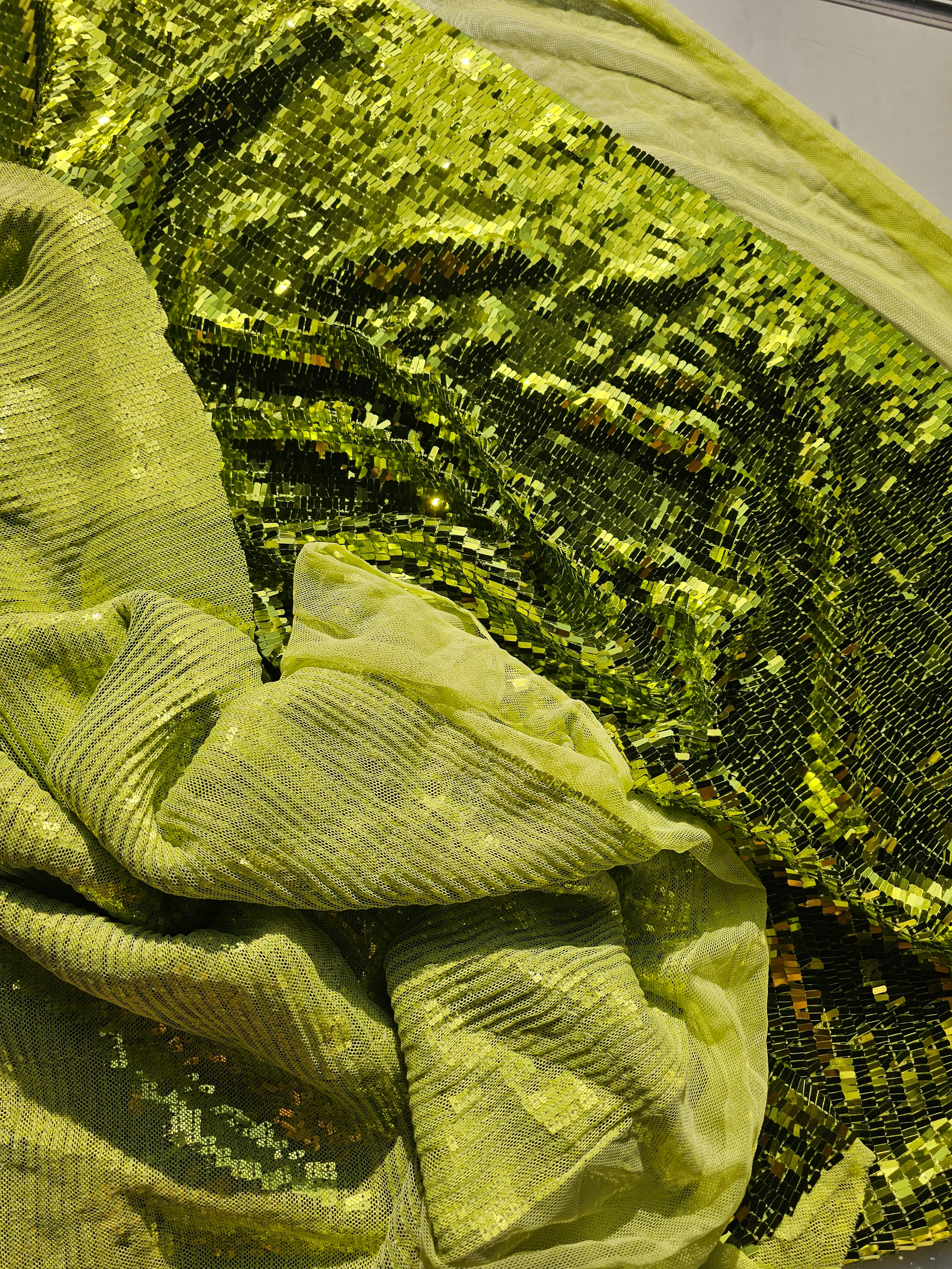 A close-up of shimmering sequined fabric in a bright chartreuse or lime green shade, with one side gathered and the other side spread out to show the reflective texture.