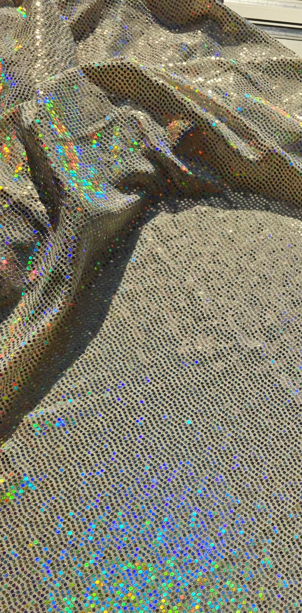 A close-up of shimmering holographic mesh fabric with a dense perforated pattern, showing iridescent rainbow reflections across a metallic taupe base.