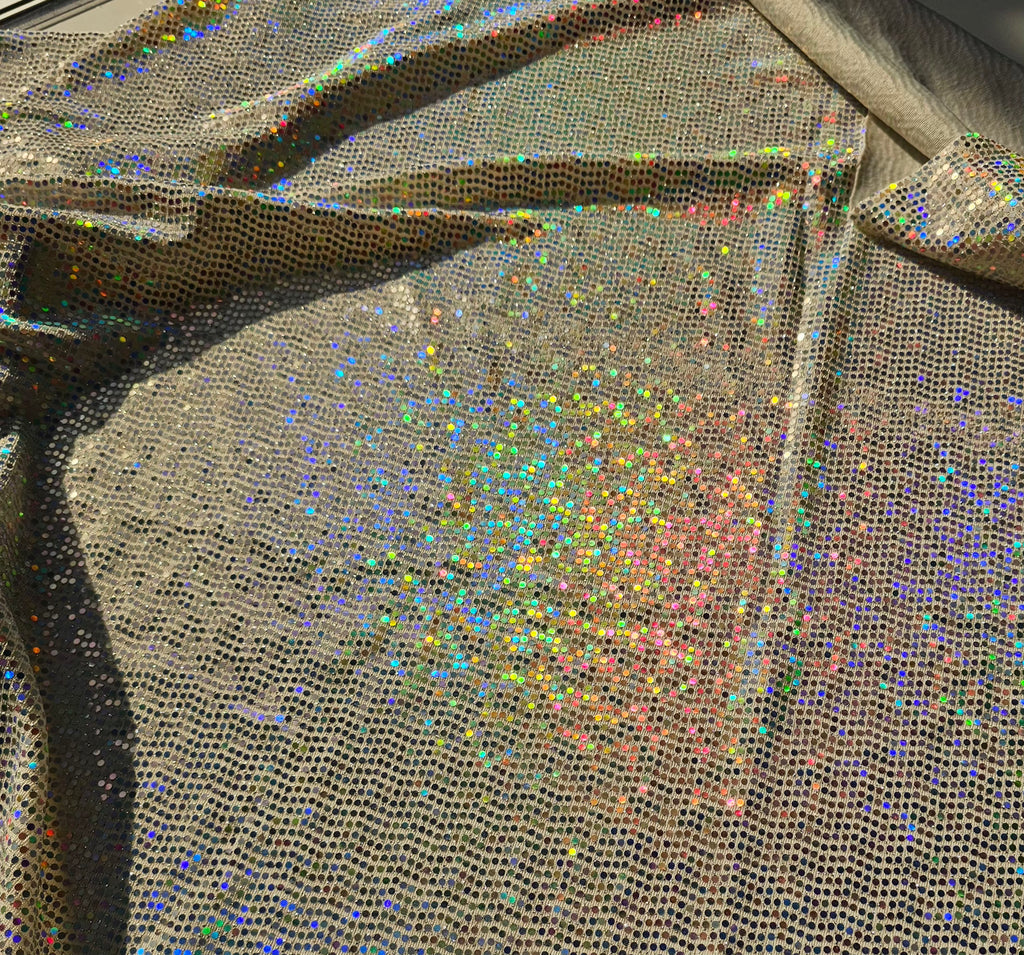Close-up of a shimmering holographic mesh fabric with dense small reflective dots in rainbow colors over a taupe-beige base, draped with visible folds and shadows.