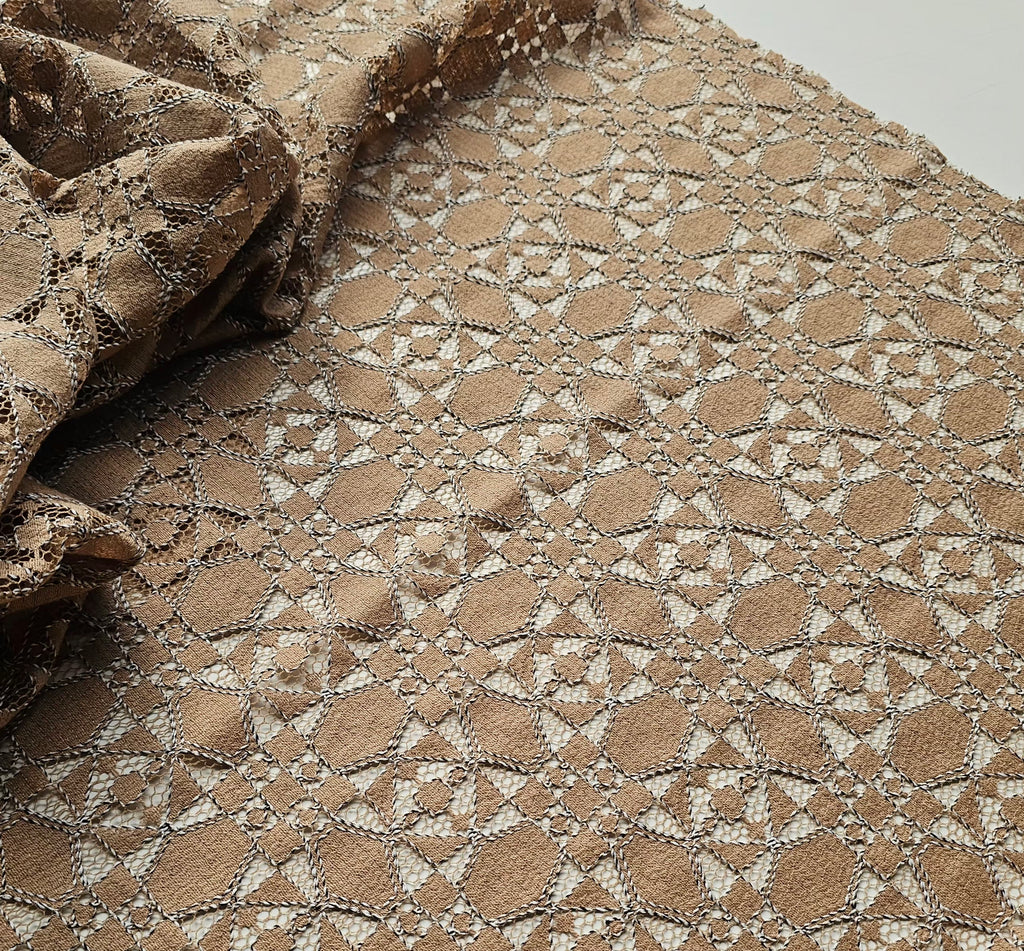 A close-up of a beige and brown lace fabric with an intricate geometric floral pattern, draped to show its texture and transparency.