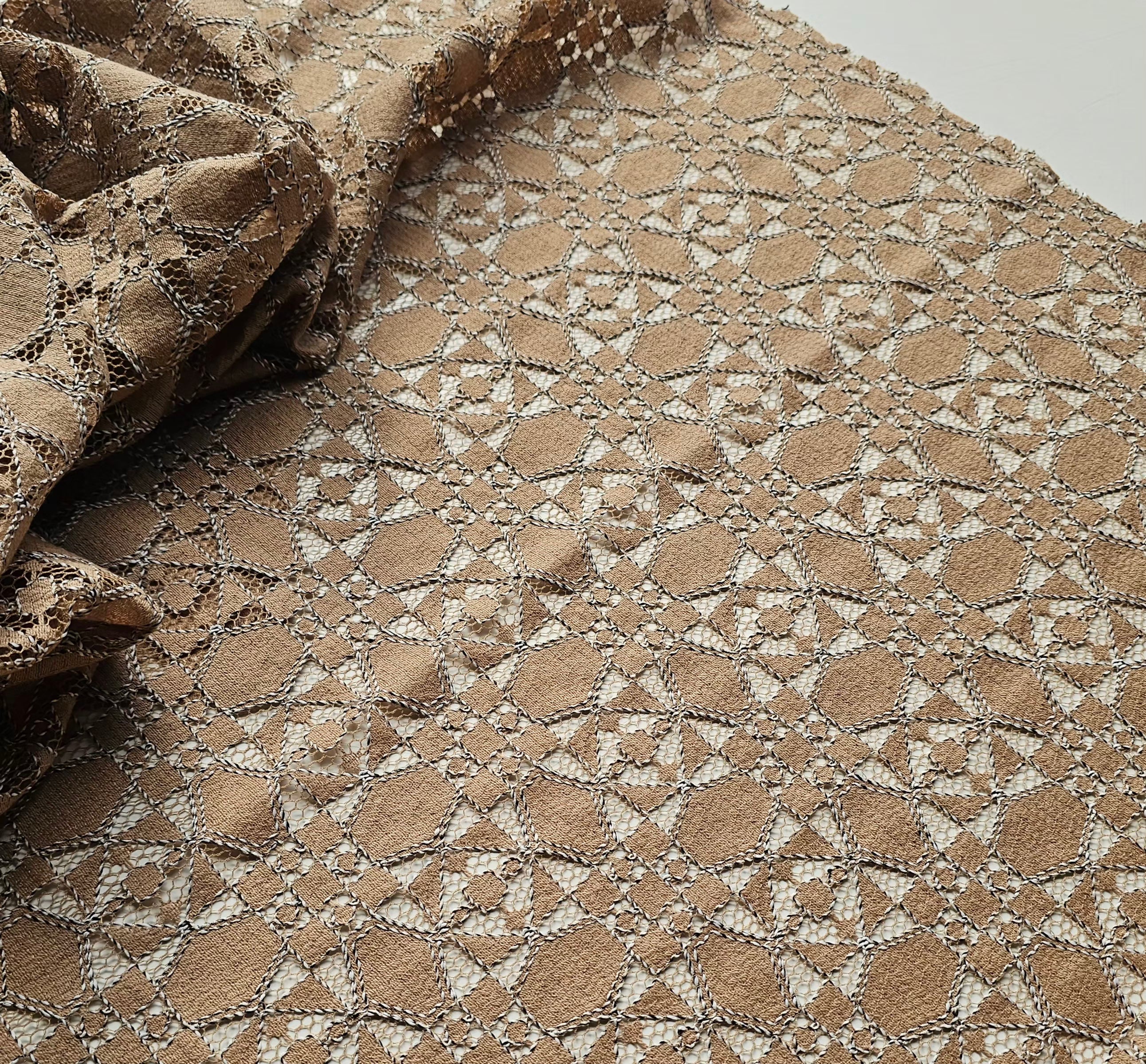 A close-up of a beige and brown lace fabric with an intricate geometric floral pattern, draped to show its texture and transparency.