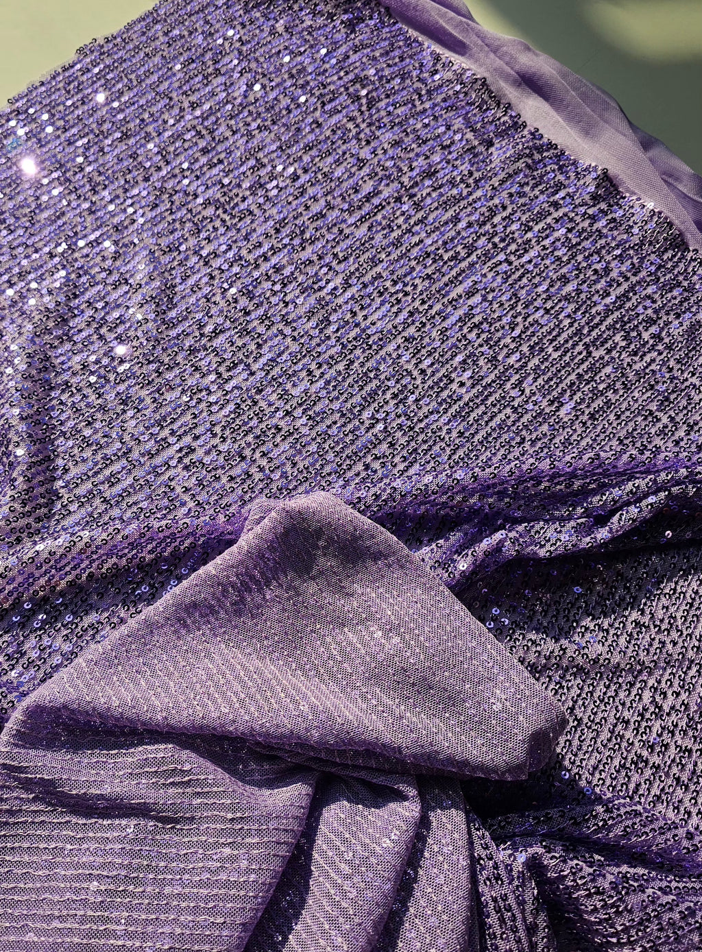 A close-up of shimmering lavender-purple sequined fabric draped and folded, showing the reflective texture and pattern of the material.
