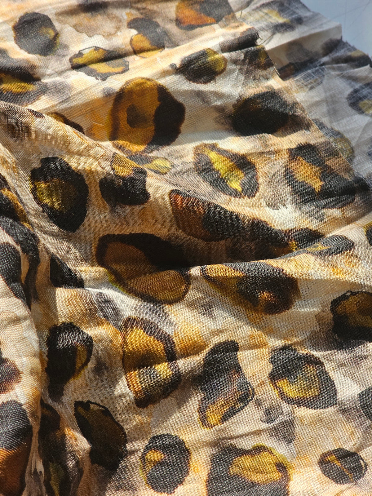 A close-up of draped fabric with an abstract leopard-like print in black, mustard yellow, and brown on a light beige background.