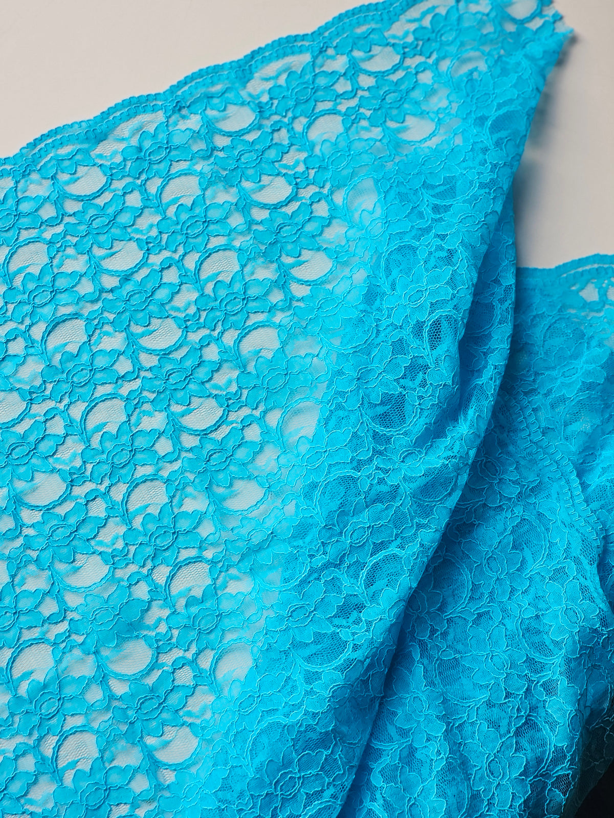 Bright turquoise blue floral lace fabric draped to show its intricate openwork pattern and scalloped edge.