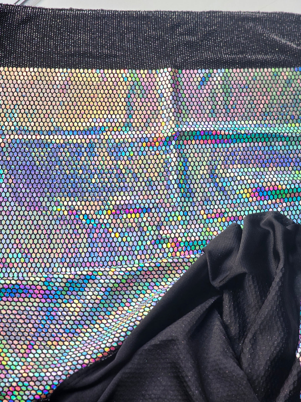 A close-up of a black mesh fabric with an iridescent holographic pattern showing rainbow reflections, with a draped black fabric overlay in the lower right corner.