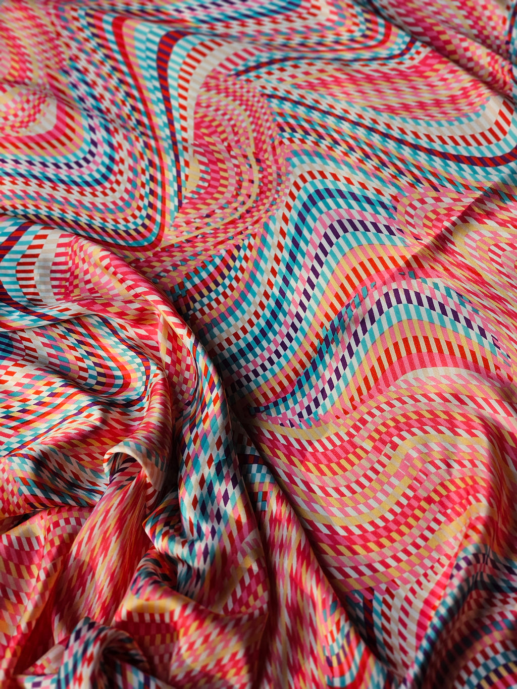 A close-up of a lightweight fabric with a vibrant, multicolor wavy pattern featuring pink, red, blue, yellow, and orange zig-zag stripes.