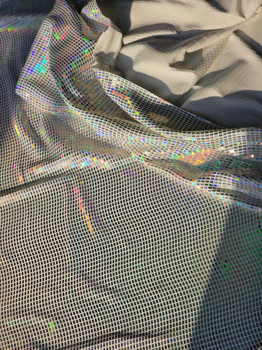 A close-up of shimmering holographic mesh fabric draped in soft folds, showing an iridescent rainbow sheen over a light gray base.