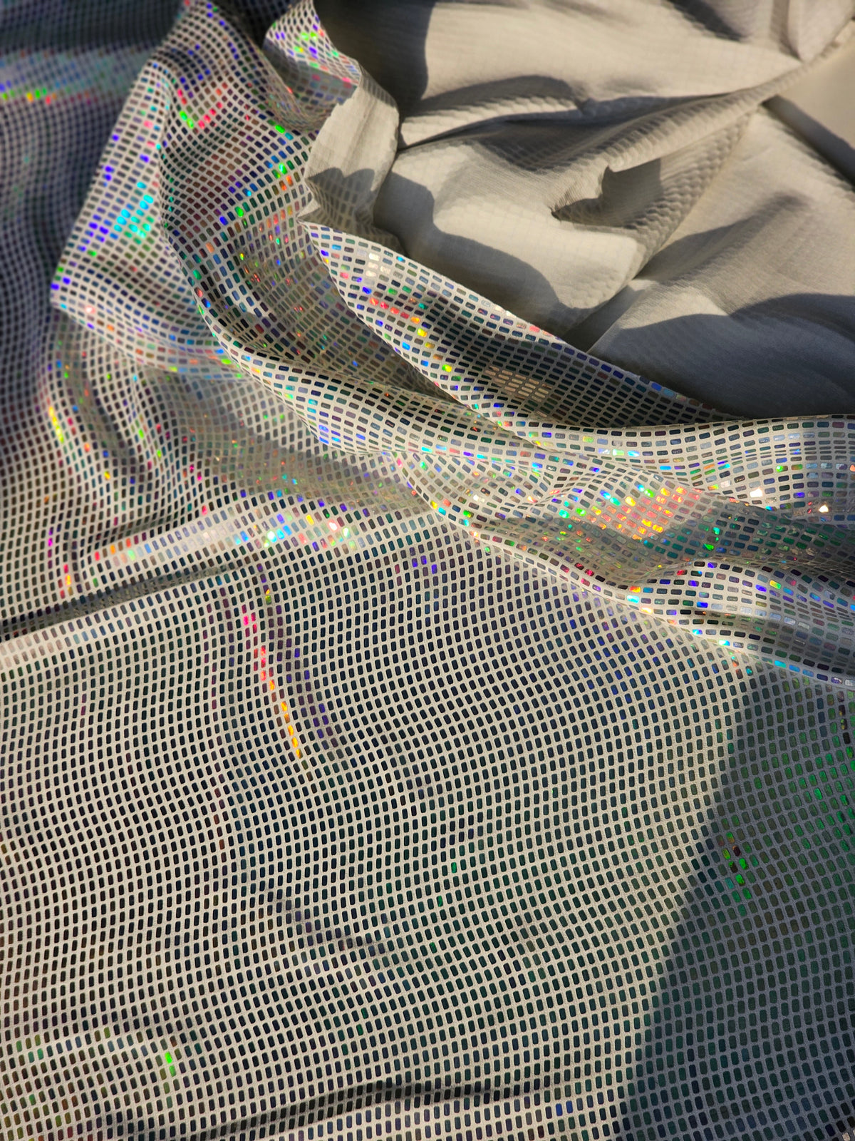 A close-up of shimmering holographic mesh fabric draped in soft folds, showing an iridescent rainbow sheen over a light gray base.