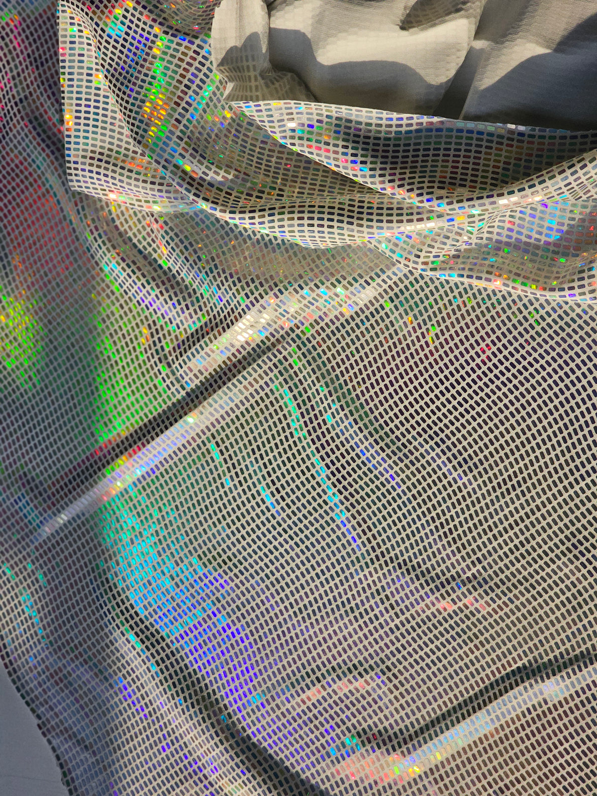 Close-up of a shimmering holographic mesh fabric with an iridescent rainbow sheen and a fine grid texture, draped in soft folds.