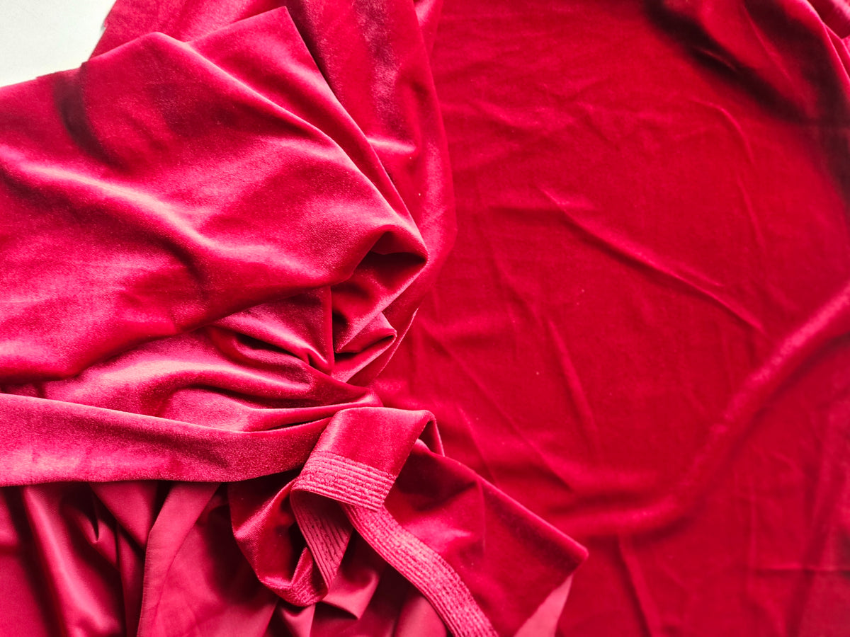 A close-up of draped red velvet fabric with soft folds and a gathered tie at the bottom left, filling the frame.
