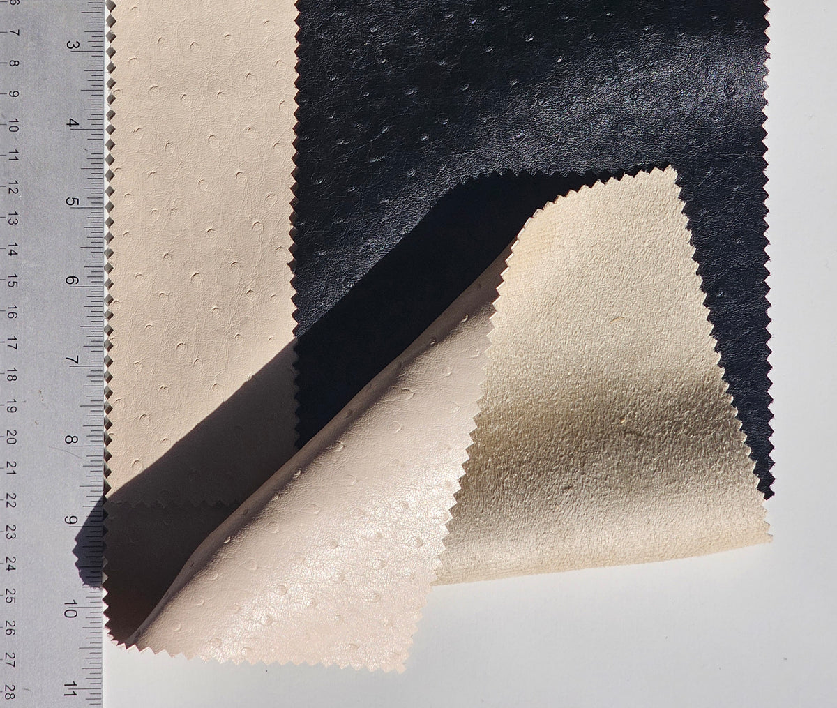 Three textured fabric swatches with scalloped edges, shown on a white background next to a ruler for scale. The fabrics are in three colors: a light beige with a dotted texture, a dark charcoal gray with a pebbled texture, and a black pebbled texture.